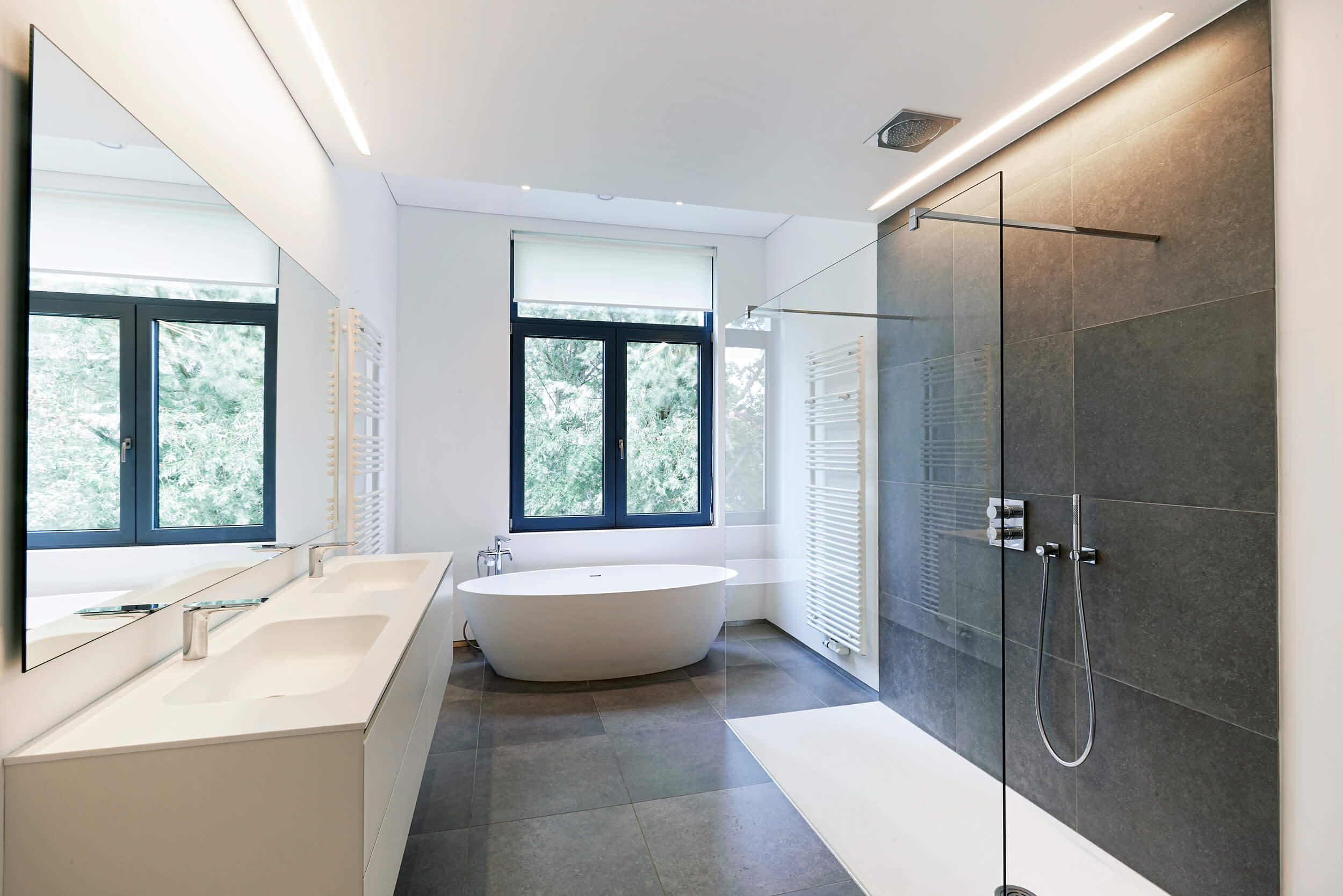 Modernes Luxus-Badezimmer mit freistehender ovaler Designerbadewanne, weißem Handtuchheizkörper und bodentiefen Fenstern. Hochwertige Badezimmerrenovierung mit grauen Bodenfliesen, weißen Wänden und eleganter Glasabtrennung - Badmodernisierung von Inotec WP in Bielefeld